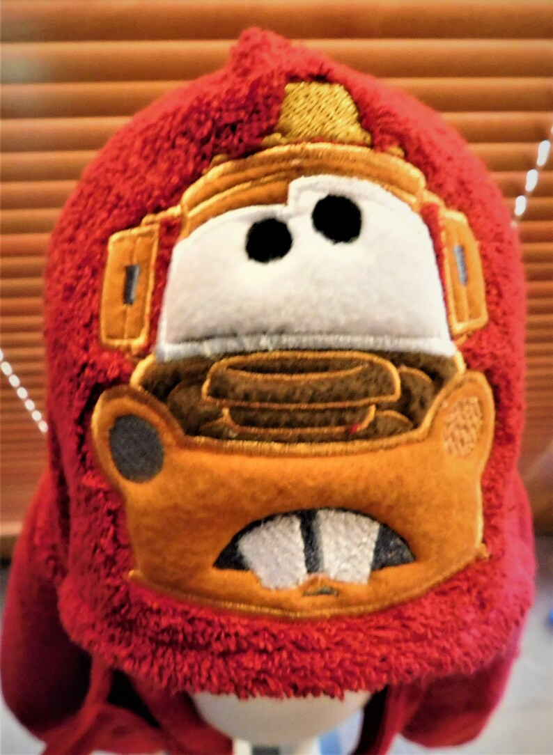 Puede incluir: Sombrero de felpa rojo con una imagen bordada de un coche de dibujos animados en marr&oacute;n, naranja y amarillo con dos ojos grandes.