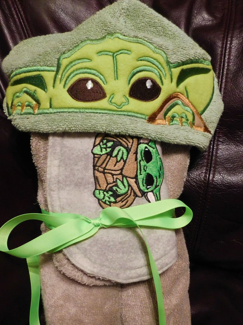 May include: A green hooded towel with an embroidered image of a character from the Star Wars franchise. The towel is tied with a green ribbon.