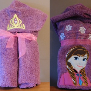 May include: Purple hooded towel with a pink ribbon and embroidered snowflake and crown designs. The towel also features an embroidered image of a girl with brown hair wearing a pink dress.