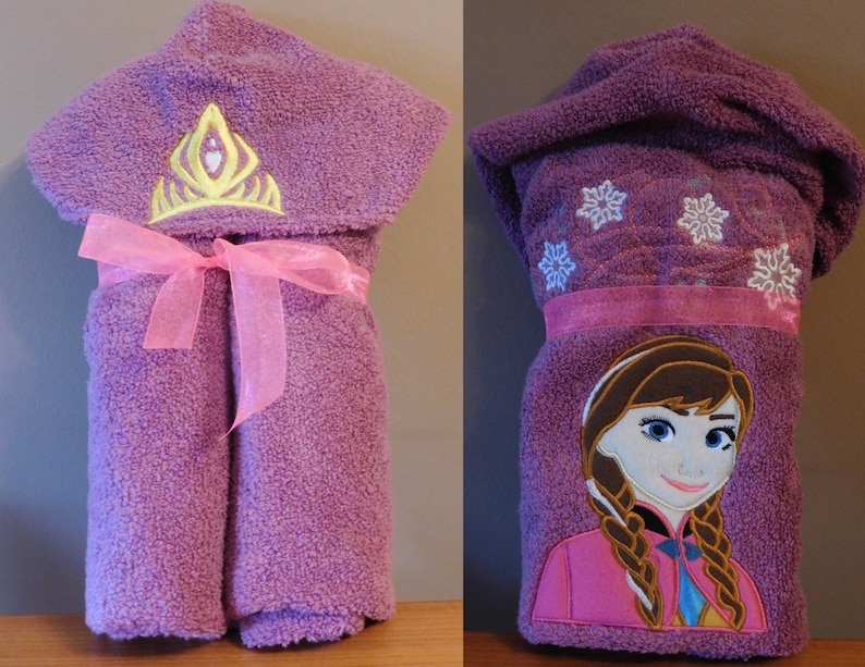 May include: Purple hooded towel with a pink ribbon and embroidered snowflake and crown designs. The towel also features an embroidered image of a girl with brown hair wearing a pink dress.