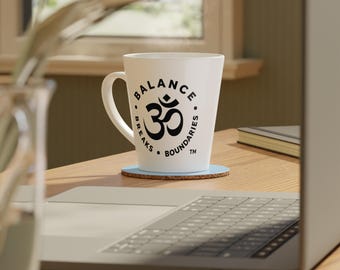 Könnte beinhalten: Ein weißer Keramik-Latte-Becher mit konischer Form und Henkel. Der Becher zeigt einen schwarzen Grafikdruck eines Om-Symbols, das von einem kreisförmigen Schriftzug mit dem Text BALANCE BREAKS BOUNDARIES TM in fetter serifenloser Schrift umgeben ist.