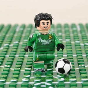 May include: A Lego figurine of a soccer goalie in a green uniform with the Emirates Fly Better logo, standing on a green field with a soccer ball. The figurine has black hair and a smiling face.