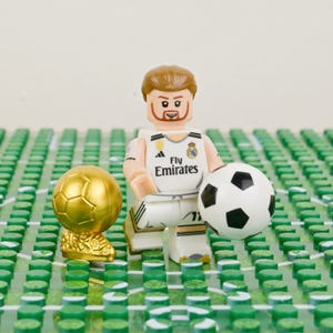 Pu&ograve; includere: Una statuina Lego di un calciatore con una maglia bianca con la scritta "Fly Emirates", che tiene un pallone da calcio. Sono presenti anche un trofeo d'oro e un pallone da calcio. La statuina &egrave; su un campo verde.