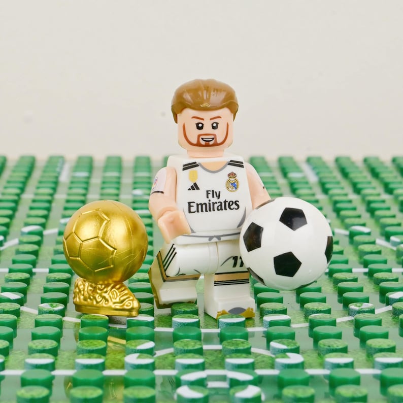 Pu&ograve; includere: Una statuina Lego di un calciatore con una maglia bianca con la scritta "Fly Emirates", che tiene un pallone da calcio. Sono presenti anche un trofeo d'oro e un pallone da calcio. La statuina &egrave; su un campo verde.
