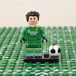 May include: A Lego minifigure of a soccer goalie in a green uniform with the text "Emirates Fly Better". A black and white soccer ball sits next to the figure on a black base. The background is a green Lego field.