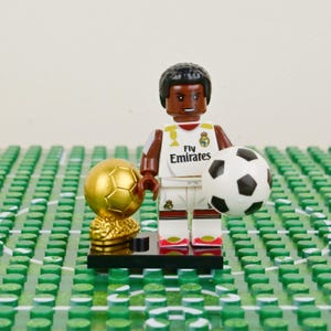 Puede incluir: Una figura de Lego de un jugador de f&uacute;tbol con uniforme blanco y marr&oacute;n, sosteniendo un bal&oacute;n de f&uacute;tbol blanco y negro. Tambi&eacute;n hay un trofeo dorado y una base verde. El uniforme tiene el texto "Fly Emirates".