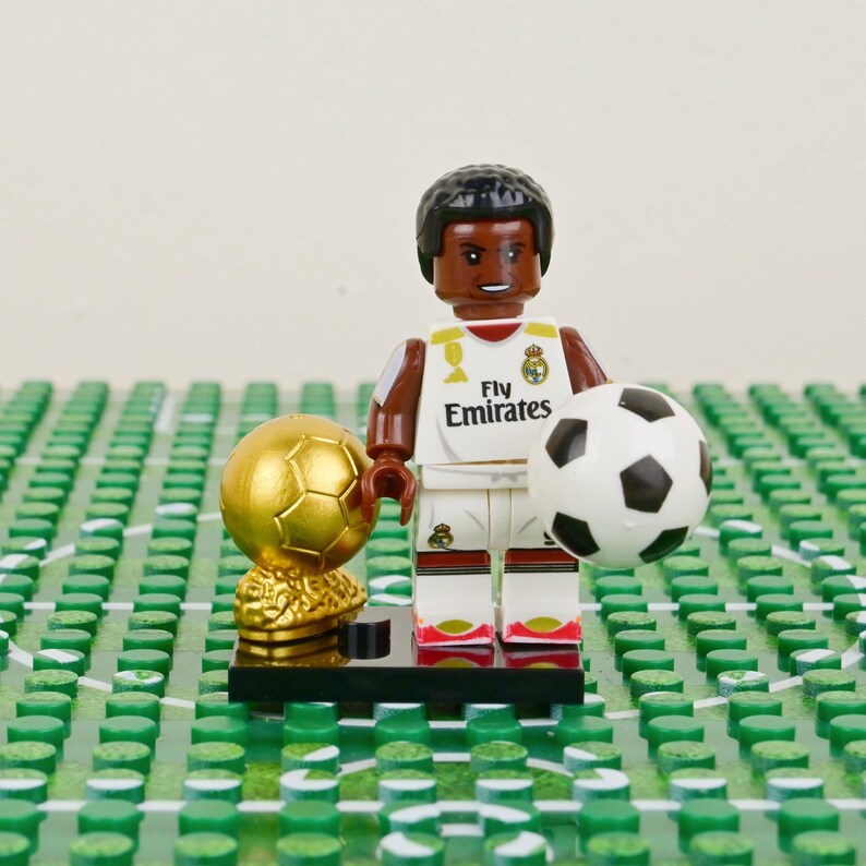 Puede incluir: Una figura de Lego de un jugador de f&uacute;tbol con uniforme blanco y marr&oacute;n, sosteniendo un bal&oacute;n de f&uacute;tbol blanco y negro. Tambi&eacute;n hay un trofeo dorado y una base verde. El uniforme tiene el texto "Fly Emirates".