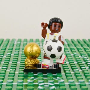 Puede incluir: Una figura de Lego de un futbolista con uniforme blanco y dorado, sosteniendo un bal&oacute;n de f&uacute;tbol blanco y negro. Un trofeo dorado est&aacute; a la izquierda. La figura est&aacute; sobre una base negra, sobre un fondo verde y blanco.
