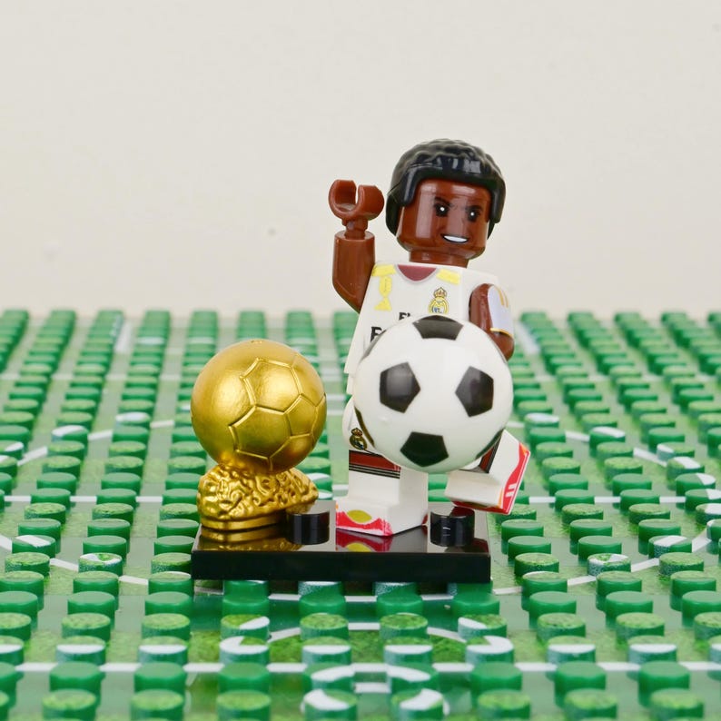 Puede incluir: Una figura de Lego de un futbolista con uniforme blanco y dorado, sosteniendo un bal&oacute;n de f&uacute;tbol blanco y negro. Un trofeo dorado est&aacute; a la izquierda. La figura est&aacute; sobre una base negra, sobre un fondo verde y blanco.