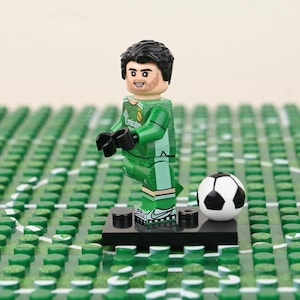 May include: A Lego figurine of a soccer goalie in a green uniform with black gloves, standing on a black base. A black and white soccer ball is next to the figurine. The background is a green surface with white lines.
