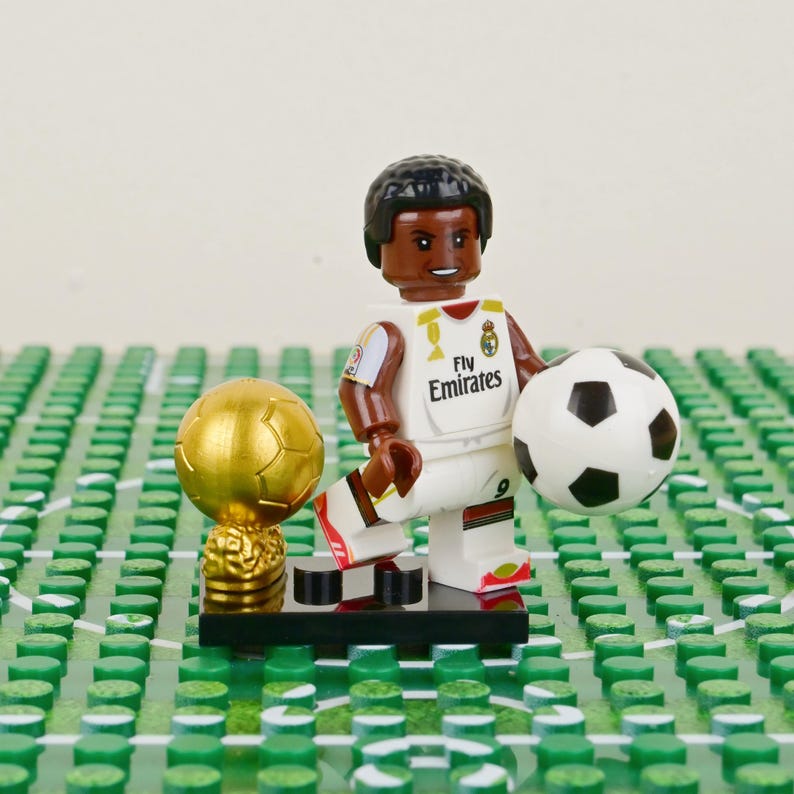 Puede incluir: Una figura de Lego de un jugador de f&uacute;tbol con uniforme blanco con el logo "Fly Emirates", sosteniendo un bal&oacute;n de f&uacute;tbol. Tambi&eacute;n hay un trofeo dorado y una base de campo de f&uacute;tbol. La figura est&aacute; sobre una base Lego verde.