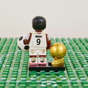 Puede incluir: Figura Lego de un futbolista con camiseta blanca con "MBAPPE" y el n&uacute;mero "9", sosteniendo un bal&oacute;n de f&uacute;tbol. Junto a &eacute;l, un trofeo dorado. La escena se desarrolla sobre un campo de Lego verde.