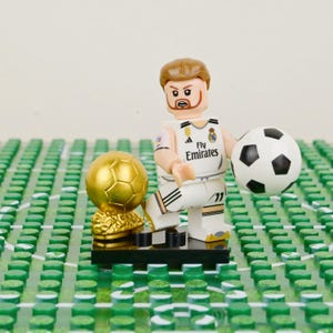 Pu&ograve; includere: Una statuina Lego di un calciatore in divisa bianca con il logo Fly Emirates, che tiene in mano un pallone da calcio bianco e nero. Un trofeo d'oro e un campo Lego verde sono anche nell'immagine.