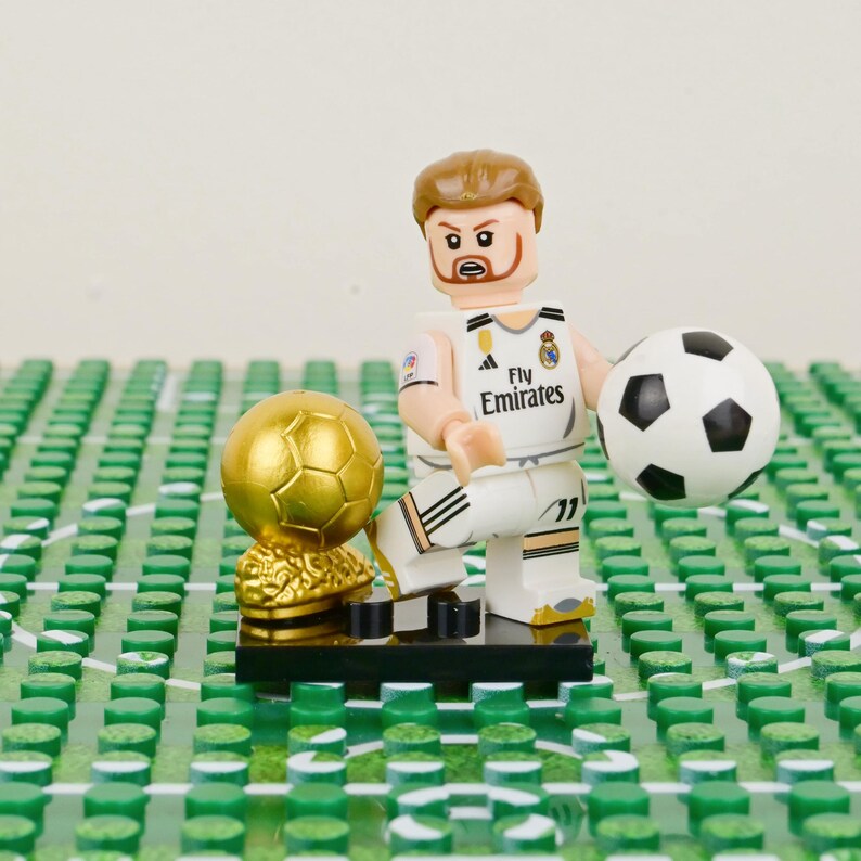 Pu&ograve; includere: Una statuina Lego di un calciatore in divisa bianca con il logo Fly Emirates, che tiene in mano un pallone da calcio bianco e nero. Un trofeo d'oro e un campo Lego verde sono anche nell'immagine.