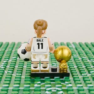 Pu&ograve; includere: Una statuina Lego di un calciatore con una maglia bianca con il nome "BALE" e il numero "11". La statuina tiene in mano un pallone da calcio e un trofeo d'oro. Si trova su un campo Lego verde.