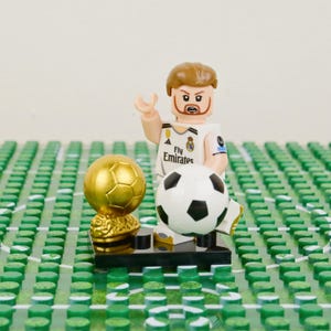 Pu&ograve; includere: Una statuetta Lego di un calciatore con una maglia bianca con il logo Fly Emirates, che tiene un pallone da calcio e accanto a un trofeo d'oro. La scena &egrave; ambientata su una base verde a forma di stadio.