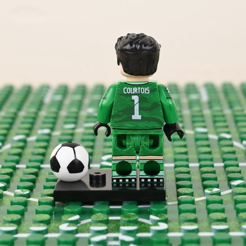 May include: A Lego figurine of a soccer goalie in a green uniform with the name "COURTOIS" and the number "1" on the back. A black and white soccer ball sits next to the figurine on a black base. The background is a green field.