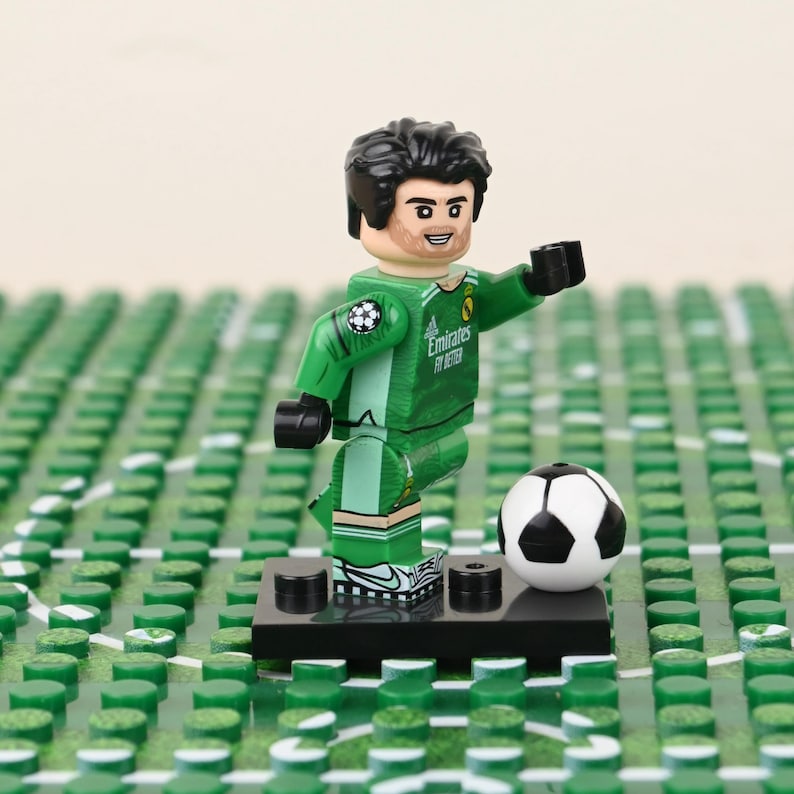 May include: A Lego soccer player figurine in a green uniform with the Emirates logo, poised to block a black and white soccer ball. The figure stands on a black base, set against a green field.