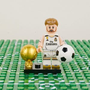 Pu&ograve; includere: Una statuina Lego di un calciatore in divisa bianca con il logo Fly Emirates, che tiene in mano un pallone da calcio bianco e nero e un trofeo d'oro. La statuina &egrave; su una base nera, su un campo verde.