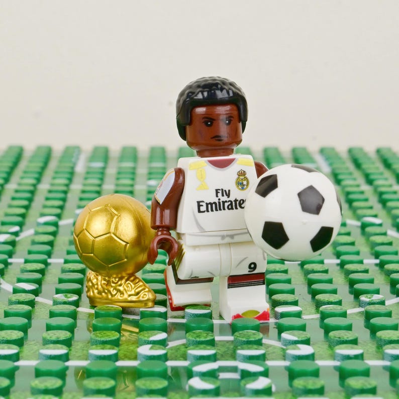 Puede incluir: Una figura de Lego de un jugador de f&uacute;tbol con uniforme blanco y marr&oacute;n con el texto "Fly Emirate" y el n&uacute;mero 9. La figura sostiene un bal&oacute;n de f&uacute;tbol blanco y negro. Un trofeo de f&uacute;tbol dorado est&aacute; a la izquierda.