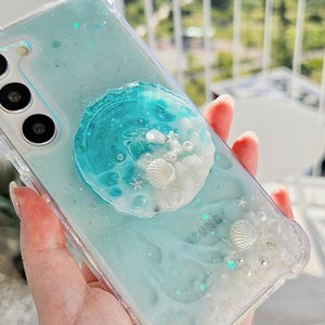 May include: A clear phone case featuring a light blue and teal resin design with embedded white seashells, small starfish, and pearl-like beads. A circular resin grip attachment with a matching ocean-inspired theme is centered on the back.