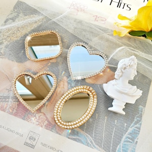 May include: A collection of four small mirrors including a rectangular mirror with a rhinestone border, two heart-shaped mirrors with rhinestone borders, and an oval mirror with a pearl border, accompanied by a small white bust sculpture.