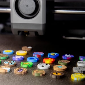 Op de afbeelding: Een verzameling kleurrijke, cirkelvormige objecten met verschillende ontwerpen en patronen. De objecten zijn gerangschikt op een donkere ondergrond, met een 3D-printer op de achtergrond. De objecten zijn ongeveer 2,5 cm in diameter.