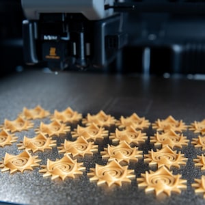 Op de afbeelding: Een 3D-printerkop boven een bouwplaat met een raster van vierentwintig kleine, lichtbruine, zonvormige plastic objecten. Elk object heeft een centraal spiraalontwerp omringd door puntige stralen, allemaal gerangschikt in uniforme rijen.
