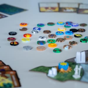 May include: A selection of brightly coloured game tokens in various designs and colours, including orange, yellow, green, blue, and brown. The tokens are scattered on a white surface, with game cards and miniature game elements in the background. The tokens appear to be made of plastic.