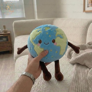 以下が含まれることがあります： 笑顔の顔、青い海、緑の大陸が特徴の地球のぬいぐるみ。茶色の腕と脚があります。おもちゃは人の手で持たれています。