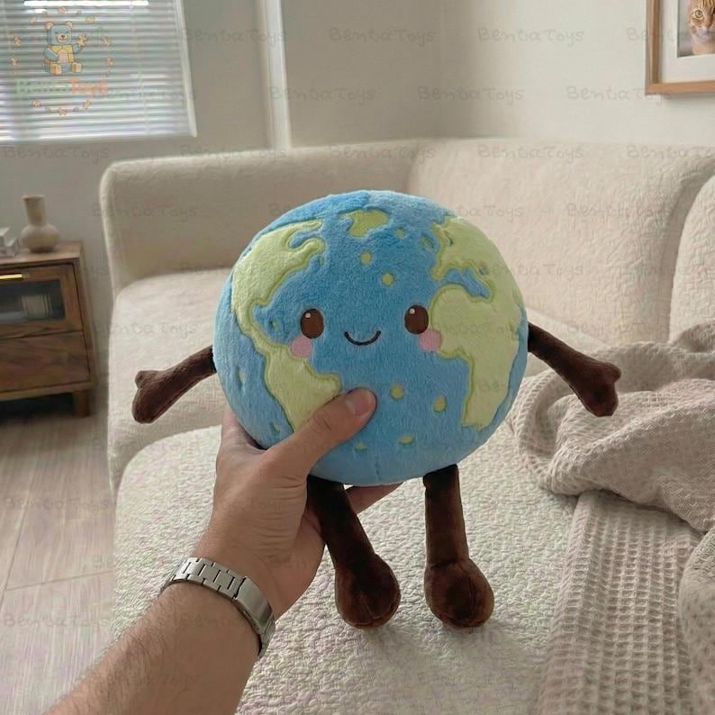 以下が含まれることがあります： 笑顔の顔、青い海、緑の大陸が特徴の地球のぬいぐるみ。茶色の腕と脚があります。おもちゃは人の手で持たれています。