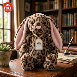 May include: A plush leopard-print bunny with long ears and a pink inner ear. The bunny has a pink nose and a tag that says "Jellycat". The bunny is sitting on a brown surface.