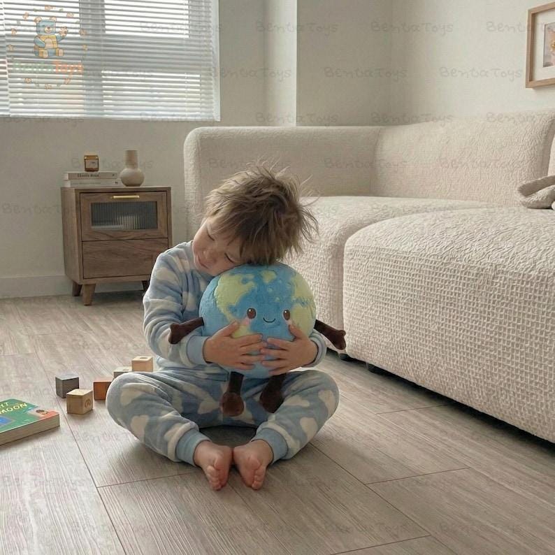 以下が含まれることがあります： 青いパジャマを着た子供が、地球のぬいぐるみと抱き合っています。地球のぬいぐるみは青と緑で、笑顔の顔と茶色の手足が付いています。子供は明るい色の木の床に座っています。