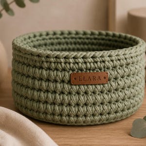 May include: A round, sage green crochet basket with a brown leather tag that reads "ELARA". The basket is made of thick yarn and has a textured appearance. The basket is sitting on a wooden surface.