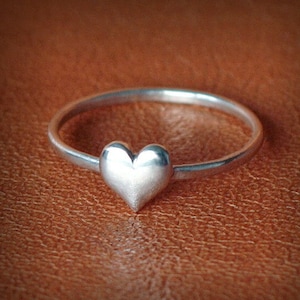 May include: A silver ring with a heart-shaped design. The ring is on a brown leather surface.