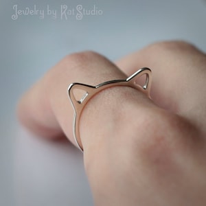 May include: A silver cat ear ring. The ring is made of a thin metal band with two cat ears on the top. The ring is worn on a person's finger.