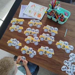 Könnte beinhalten: Ein Set aus gelben Muldenkipper-Ausschnitten mit mathematischen Additionsaufgaben und den dazugehörigen Ergebnissen. Die Lkws sind auf einer Fläche angeordnet, neben einem hellblauen Kunststoff-Organizer mit Buntstiften und einem Heft namens Explode The Code.