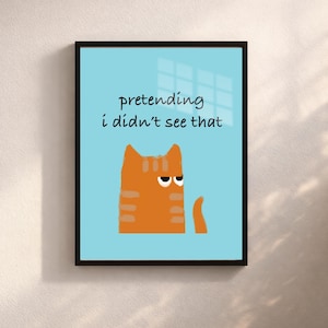 Puede incluir: Una impresión artística enmarcada con un gato naranja de dibujos animados sobre un fondo azul claro. El gato tiene una expresión gruñona. El texto "pretending i didn't see that" está encima del gato. La obra de arte está en un marco negro.