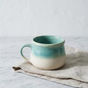 May include: A handmade ceramic mug with a rounded base and a small handle. The mug features a two-tone design, with a light teal glaze at the top that fades into a creamy white at the bottom. The mug is sitting on a beige linen napkin.