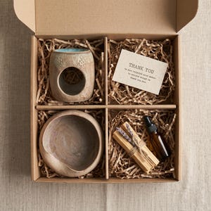 Può includere: Una scatola regalo in cartone contenente un bruciatore di oli essenziali in ceramica con interno blu, una ciotola rotonda in ceramica, un fascio di legnetti di palo santo legati con spago e lavanda, un flacone contagocce in vetro ambrato e una carta THANK YOU.