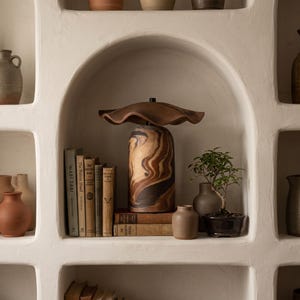 Puede incluir: Una lámpara de mesa con base cilíndrica que presenta un patrón jaspeado en tonos marrón, beige y negro. La lámpara tiene una pantalla ancha, ondulada y de color marrón oscuro. Se sitúa sobre libros junto a un bonsái en un estante blanco.