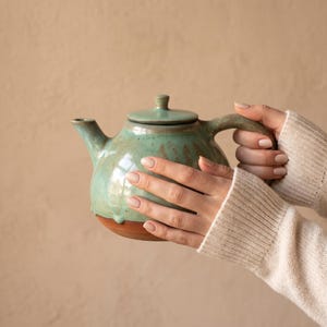 Könnte beinhalten: Eine Keramik-Teekanne mit bauchigem Körper und Deckel, die eine glänzende hellgrüne Glasur im oberen Bereich und eine kontrastierende, unglasierte terrakottafarbene Basis aufweist. Die Teekanne hat einen geschwungenen Ausguss und einen Griff.