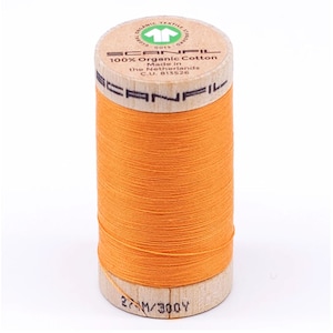 May include: A spool of bright orange Scanfil 100% Organic Cotton thread. The wooden spool has text and a green logo. The thread is tightly wound, and the spool is labeled with the text "27-M/300Y".