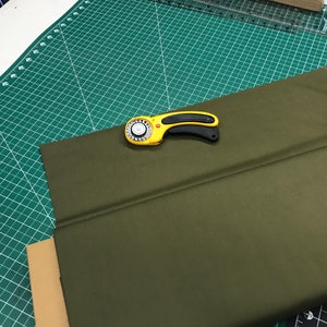May include: A green fabric is laid out on a green cutting mat with a rotary cutter on top. The fabric is folded in half.