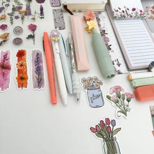 May include: An assortment of stationery items, including pens, highlighters, stickers, and a notebook. The pens are in coral, white, and light blue. Stickers feature floral designs and illustrations of flowers in jars. A notebook with the text "FLOWER & TREE" is also visible.
