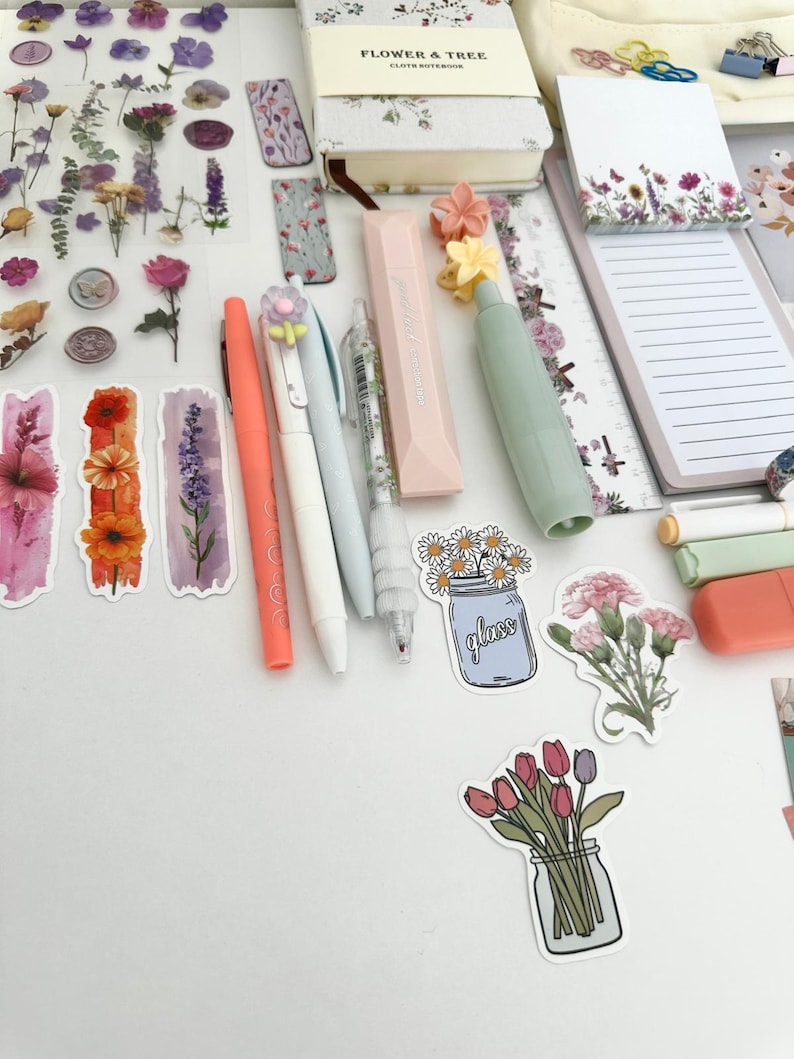 May include: An assortment of stationery items, including pens, highlighters, stickers, and a notebook. The pens are in coral, white, and light blue. Stickers feature floral designs and illustrations of flowers in jars. A notebook with the text "FLOWER & TREE" is also visible.