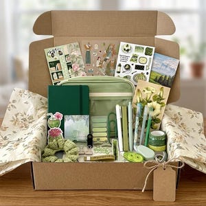 Könnte beinhalten: Eine Pappschachtel gefüllt mit Schreibwaren und Accessoires in Grüntönen. Enthalten sind ein dunkelgrünes Notizbuch, ein hellgrünes Reißverschlussetui, Stifte, Washi-Tape, Aufkleber, ein gehäkeltes Haargummi, Büroklammern und florales Seidenpapier.