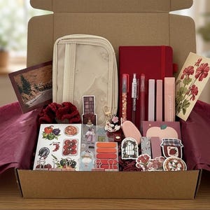 Op de afbeelding: Een cadeauverpakking gevuld met briefpapier en schoonheidsproducten. Inclusief een beige etui, een rood notitieboek, pennen, lippenstiften, stickers en een scrunchie. De doos is gevoerd met bordeauxrood vloeipapier.