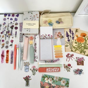May include: A collection of stationery items including a floral notebook, pens, stickers, highlighters, and a pencil case. The items are arranged on a white surface, featuring a variety of floral designs and pastel colors. The pencil case is beige and the stickers have dried flowers.
