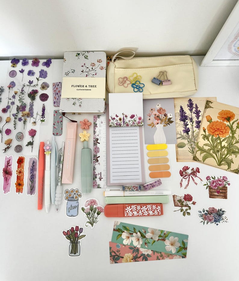 May include: A collection of stationery items including a floral notebook, pens, stickers, highlighters, and a pencil case. The items are arranged on a white surface, featuring a variety of floral designs and pastel colors. The pencil case is beige and the stickers have dried flowers.